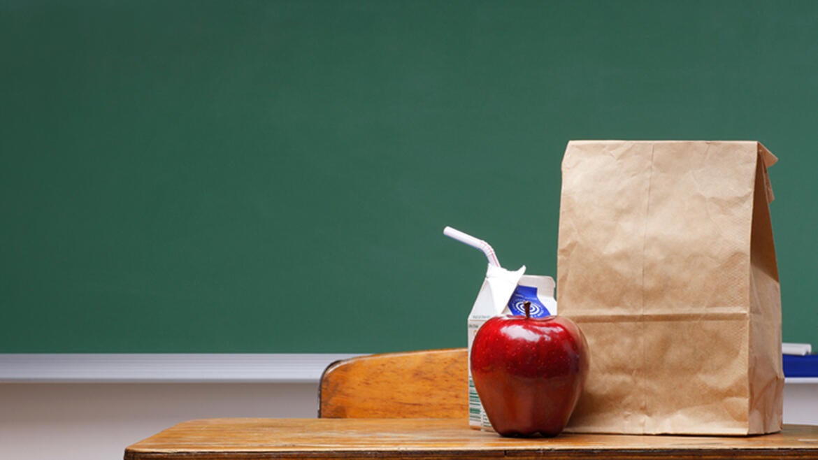 An apple, milk, and brown bag lunch on a school desk in front of a green chalkboard