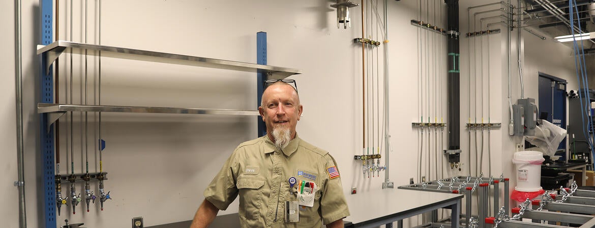 A technician standing inside a clean laboratory with overhead piping and wall-mounted valves