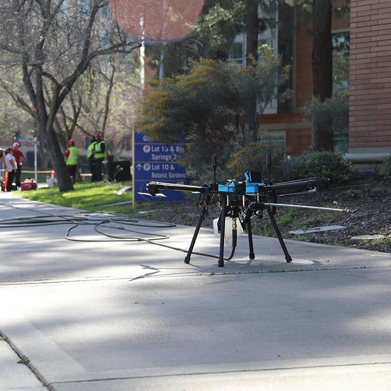 A black drone sits on the sidewalk next to a brick building with a van in the background