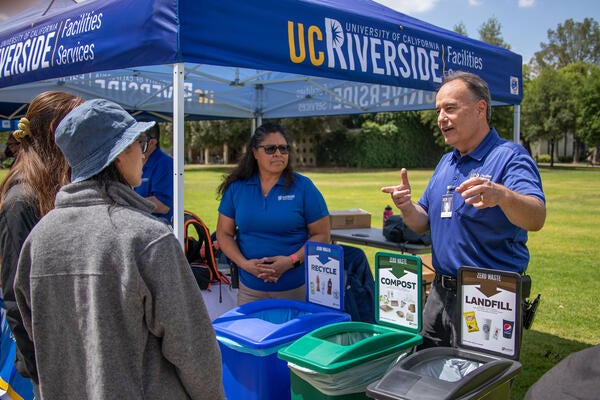 People at an outdoor booth showcasing recycling, composting, and trash bins