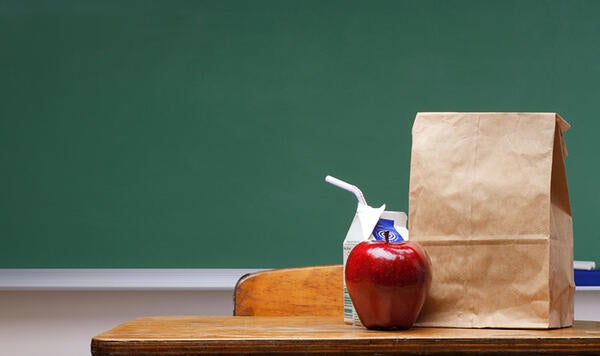 An apple, milk, and brown bag lunch on a school desk in front of a green chalkboard