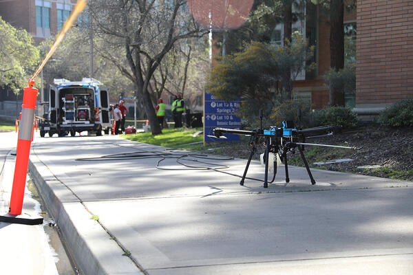A black drone sits on the sidewalk next to a brick building with a van in the background