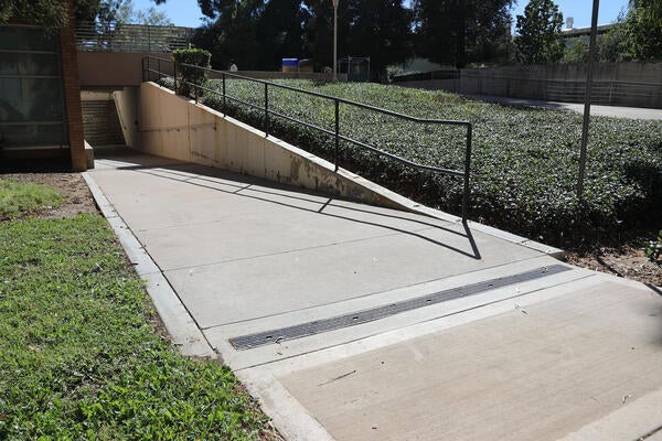 A concrete accessibility ramp with black metal handrails, sloped walkway, retaining walls, drainage grates