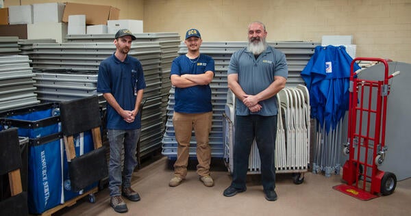 Three people standing in front of stacks of tables, chairs, and moving dollies