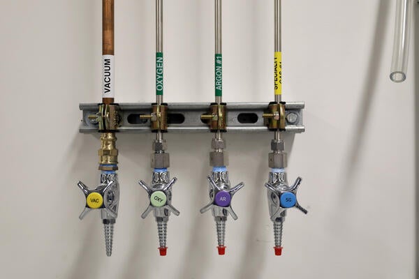 A wall-mounted laboratory gas manifold distributing vacuum, oxygen, argon, and nitrogen through four labeled, color-coated lines