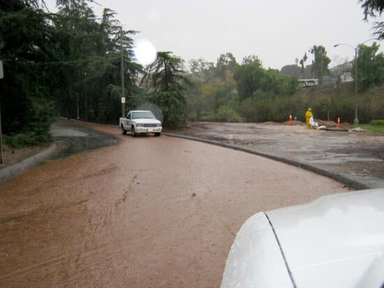 A rain-flooded road, with muddy water covering pavement and a car and worker on the right